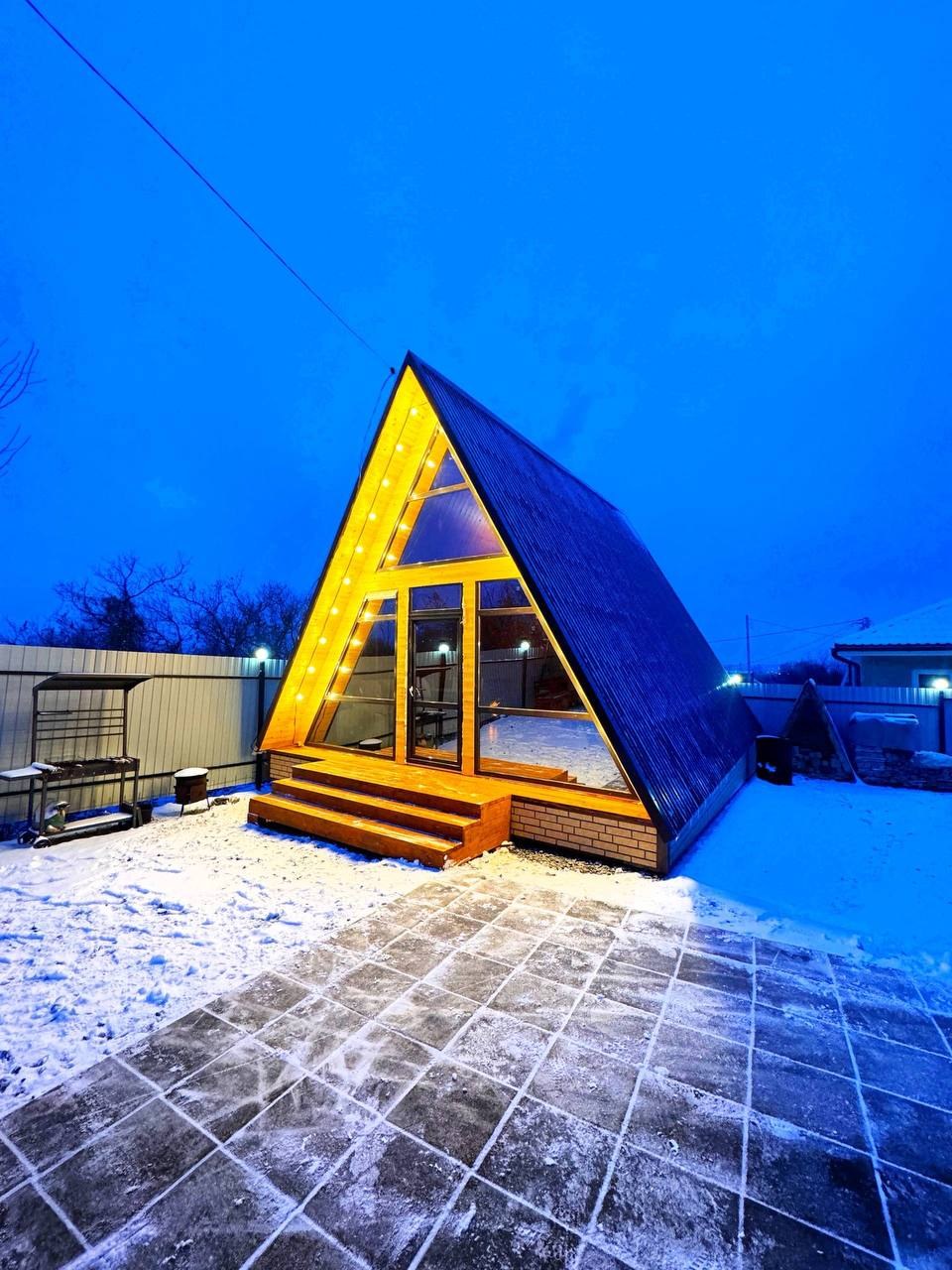 Наш дом A-Frame вечером: остеклённый фронтон с подсветкой и деревянное крыльцо